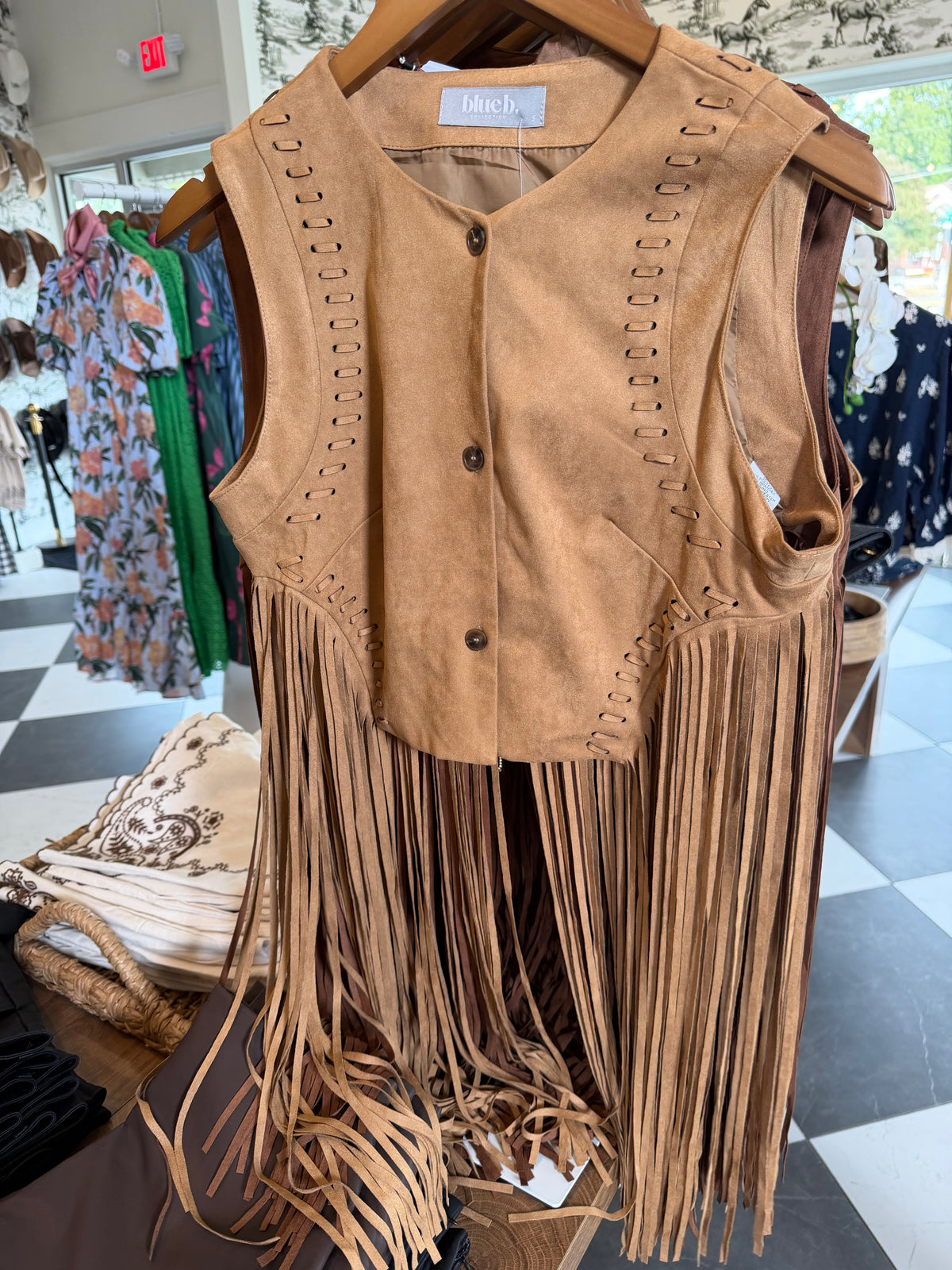 A tan suede fringe vest with decorative stitching and button-front closure, hanging on a wooden hanger inside a clothing store.
