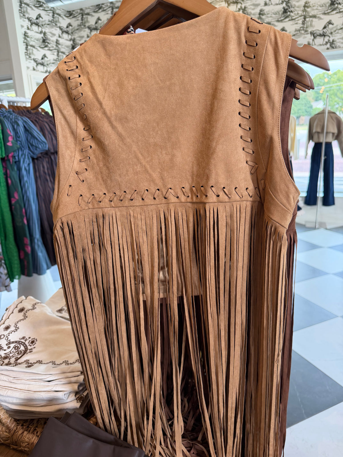 A tan suede fringe vest hanging on a wooden hanger in a clothing store. The sleeveless vest features decorative side lacing and a long fringe hem.