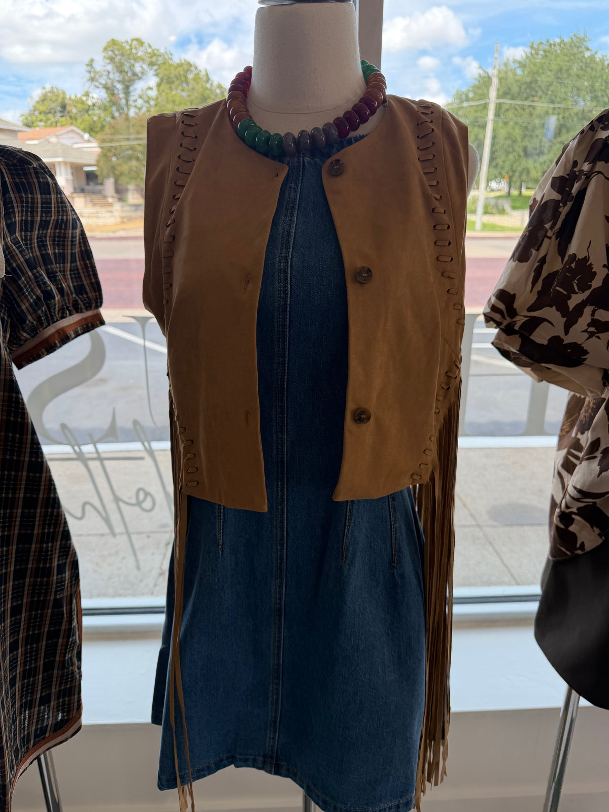 A tan suede fringe vest with button details, styled on a mannequin over a blue denim dress, with a colorful beaded necklace, displayed in a store window.