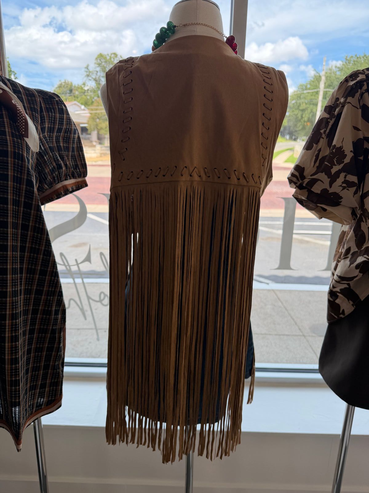 A tan faux-suede fringe vest displayed on a mannequin torso, shown from the back with decorative cutout stitching. The vest is flanked by other garments on mannequins in front of a large window showing a street scene.