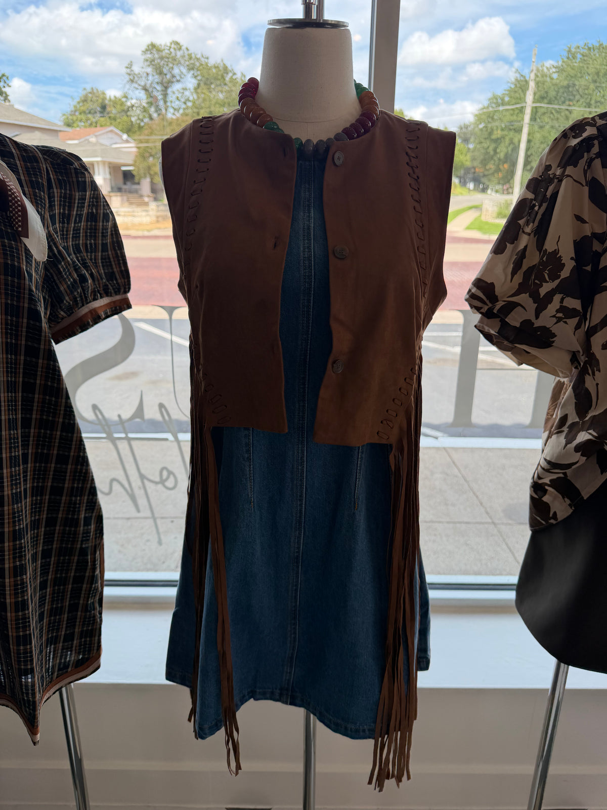 A brown sleeveless fringe vest displayed on a mannequin in a store window, next to a patterned blouse on another mannequin.
