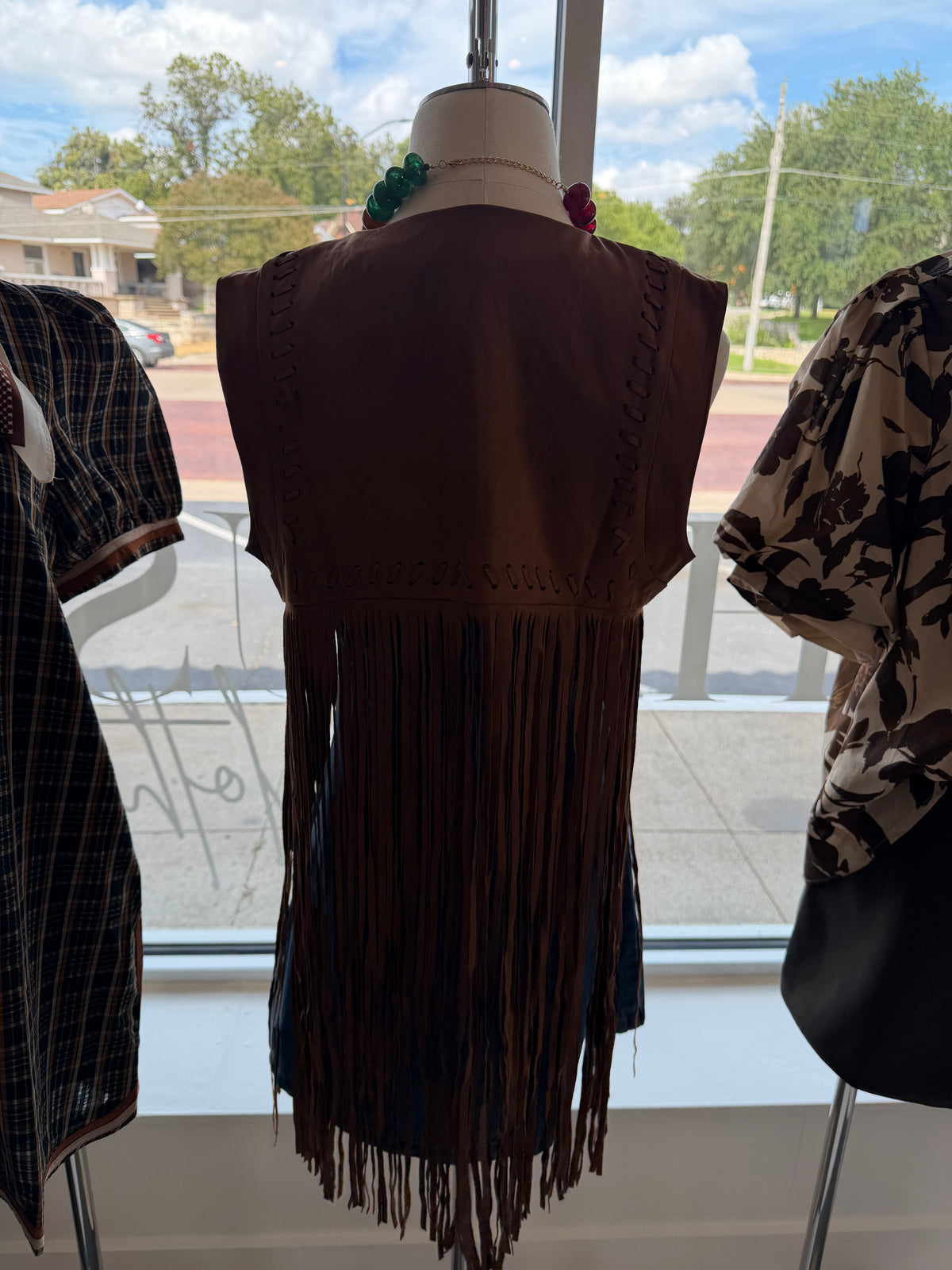 A brown sleeveless fringe vest displayed on a mannequin in a store window, next to a patterned blouse on another mannequin.