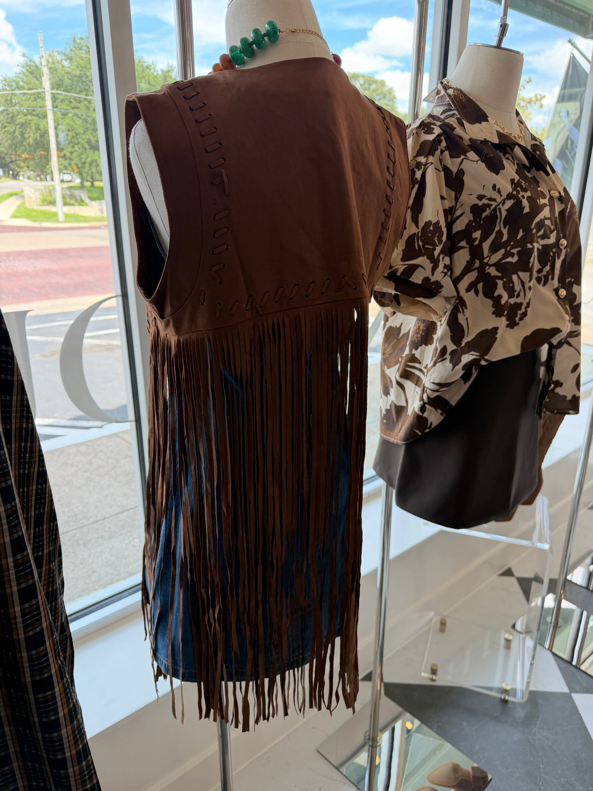 A brown sleeveless fringe vest displayed on a mannequin in a store window, next to a patterned blouse on another mannequin.