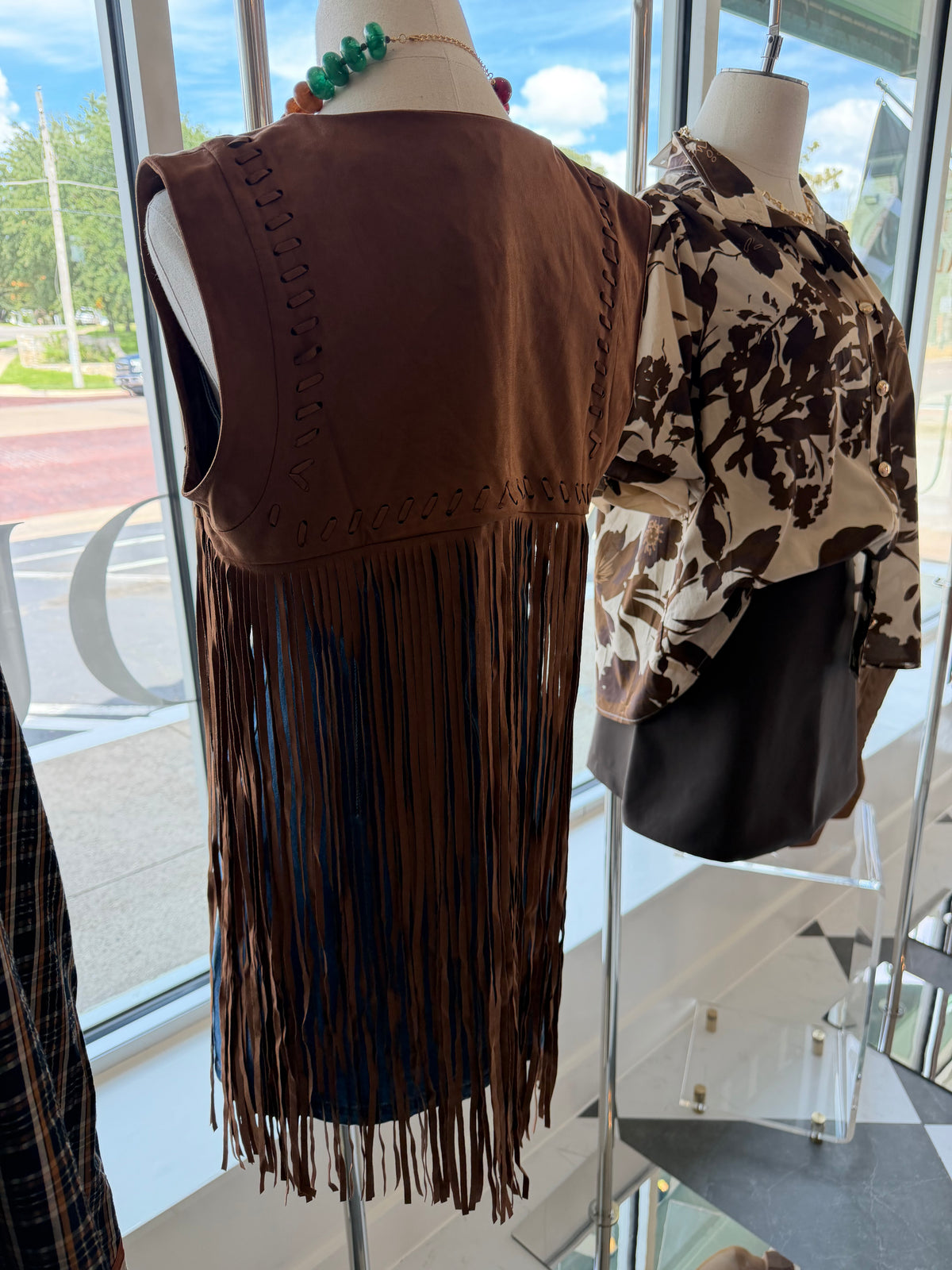 A brown faux-leather fringe vest displayed on a mannequin in a boutique window display, next to a patterned blouse and black bag.