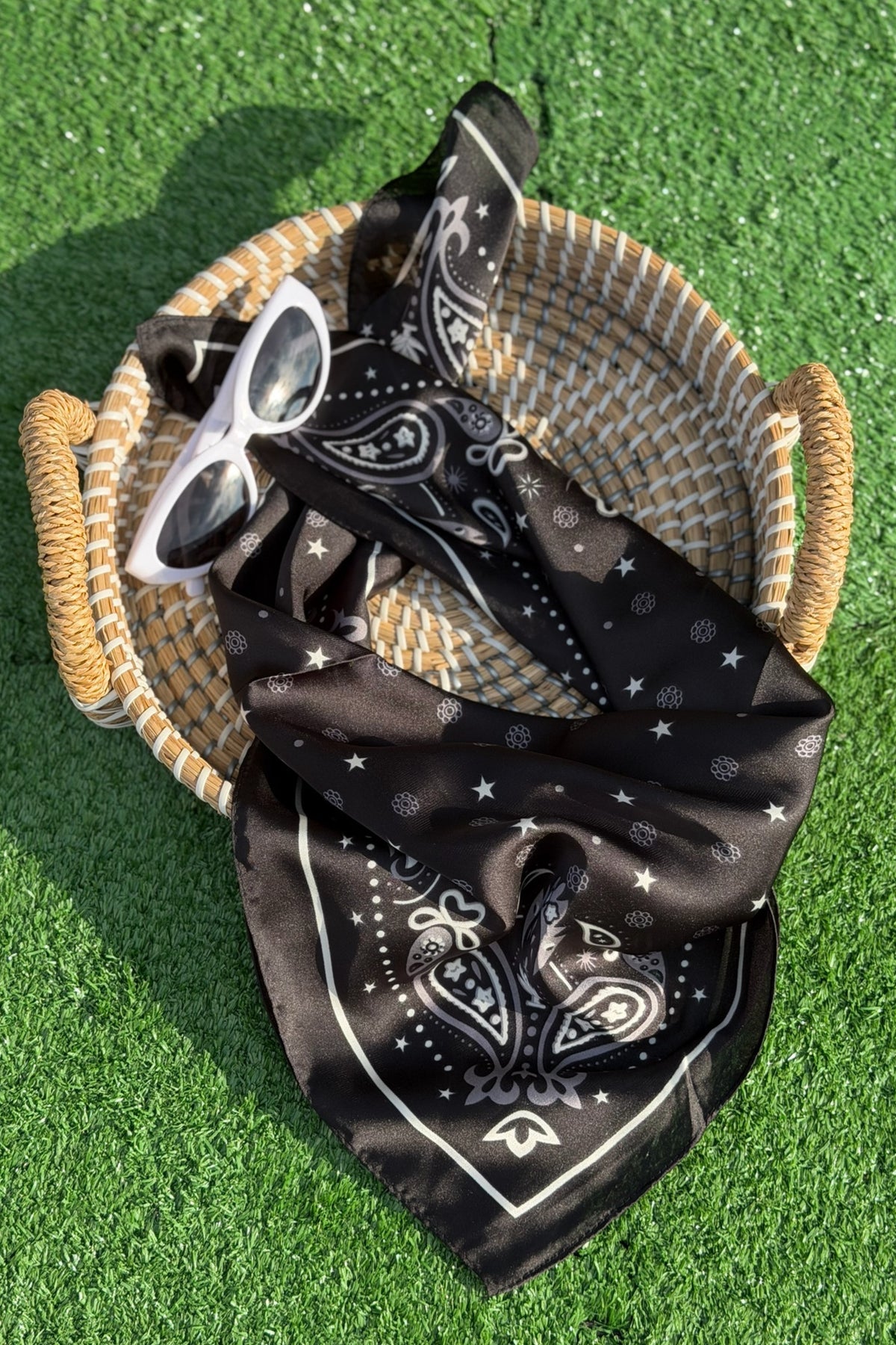Black bandana with white patterns on a woven basket with sunglasses, on a green grass background