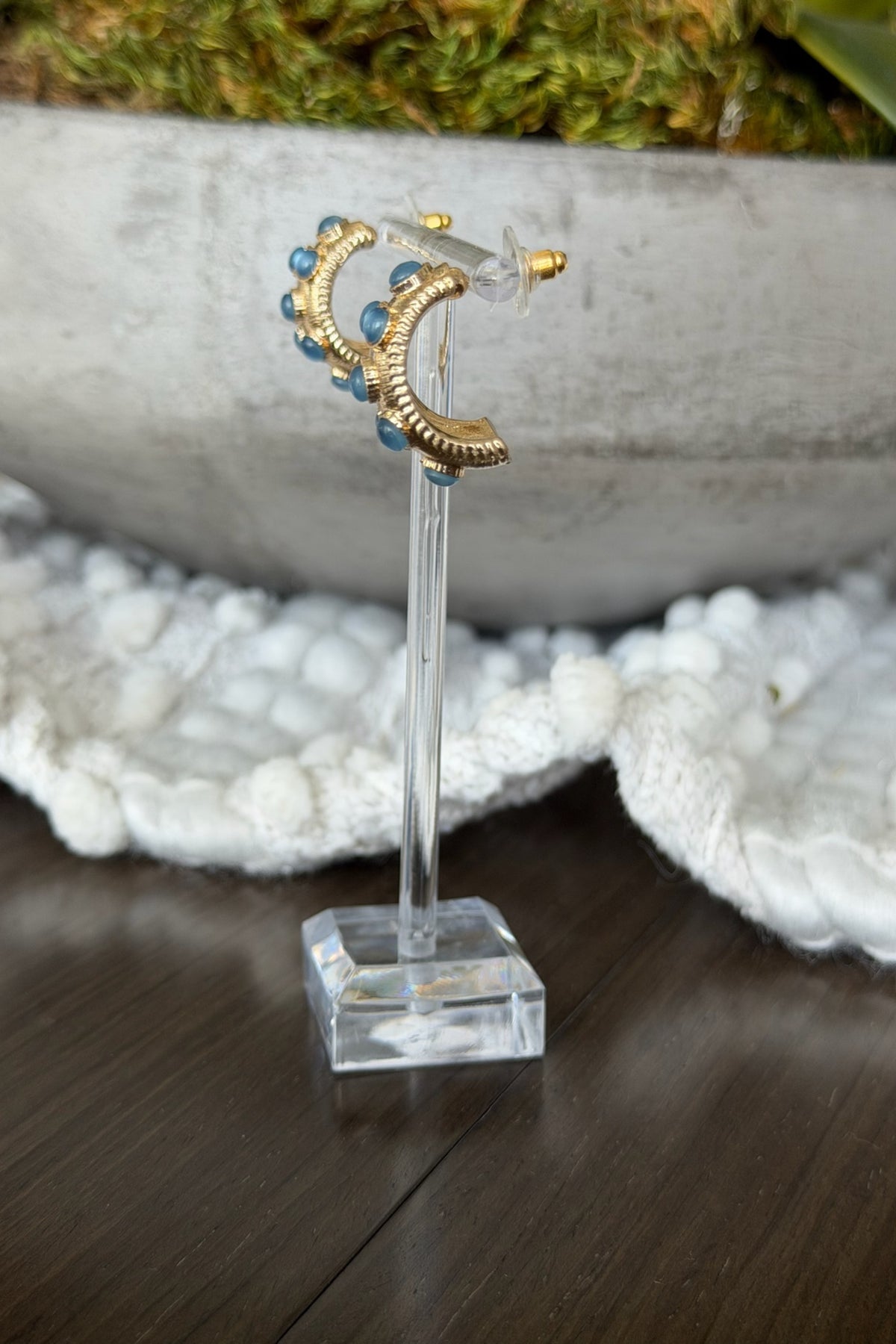 Gold moon-shaped earrings with blue stones on a clear stand against a natural background.