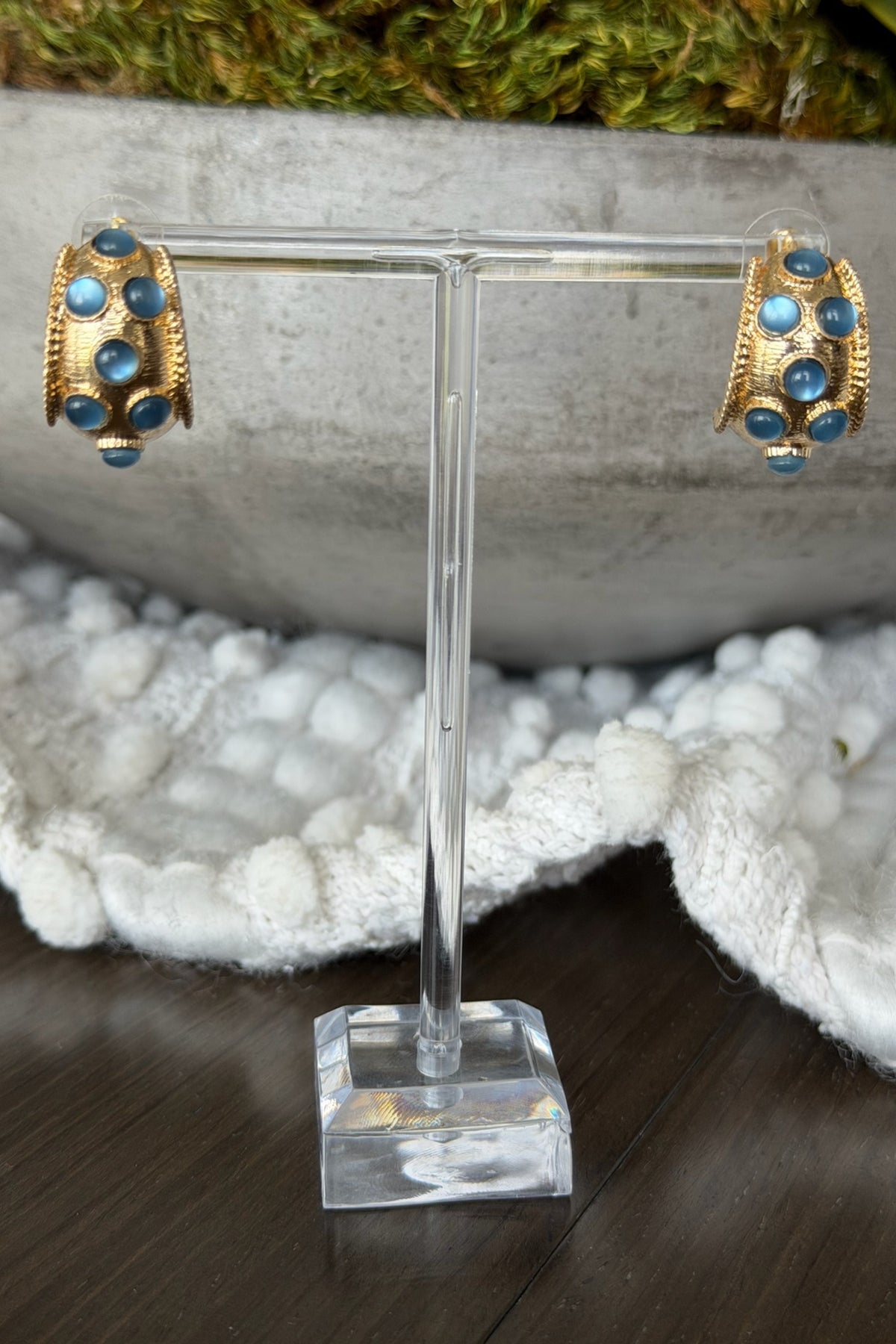 Gold earrings with blue stones on a clear stand against a natural background