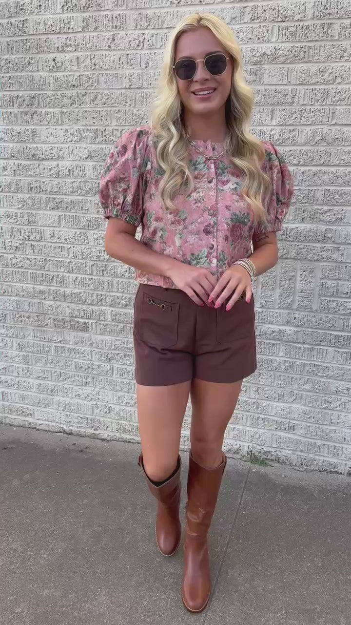 A woman wearing a pink floral puff-sleeve button-up top and brown shorts, standing outdoors on a paved walkway.