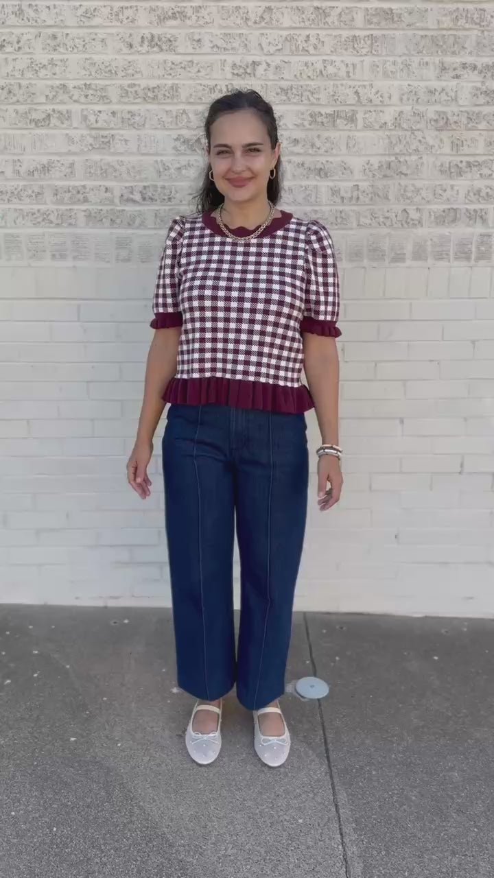A woman stands on a sidewalk wearing a short-sleeve gingham top with dark red ruffled trim and dark blue wide-leg 