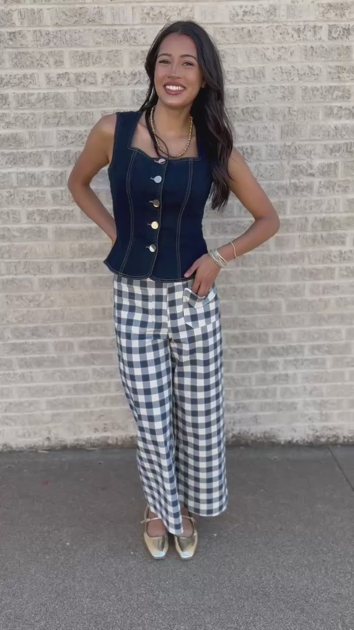 A woman stands outdoors against a light brick wall wearing a sleeveless navy top with gold buttons and blue-and-white gingham cropped pants.