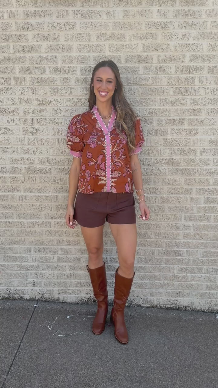 A woman walking on a sidewalk wearing a rust-orange and pink floral short-sleeve button-up shirt, brown shorts, and brown knee-high boots.