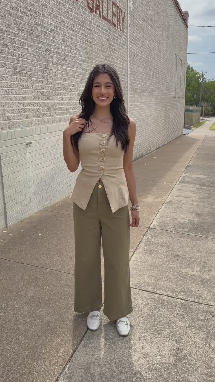 Brunette model wearing the Westlyn Buckle Strapless Top in Taupe featuring gold-tone buckle accents and a split-front hem, styled with olive wide-leg pants and white loafers standing outside. Southern House Clothing.