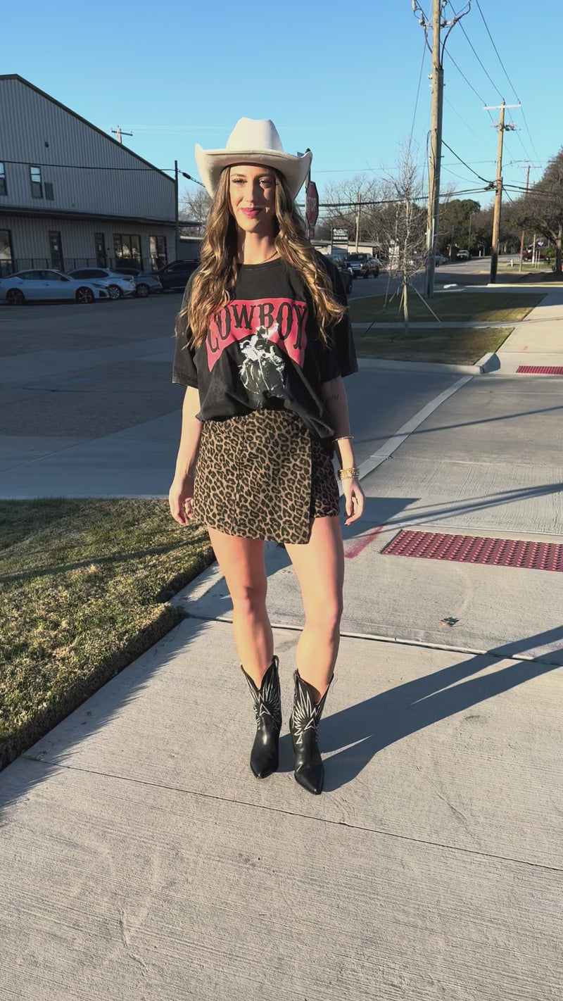 Woman wearing a cowboy graphic t-shirt, leopard print skirt, and black cowboy boots in a parking lot.