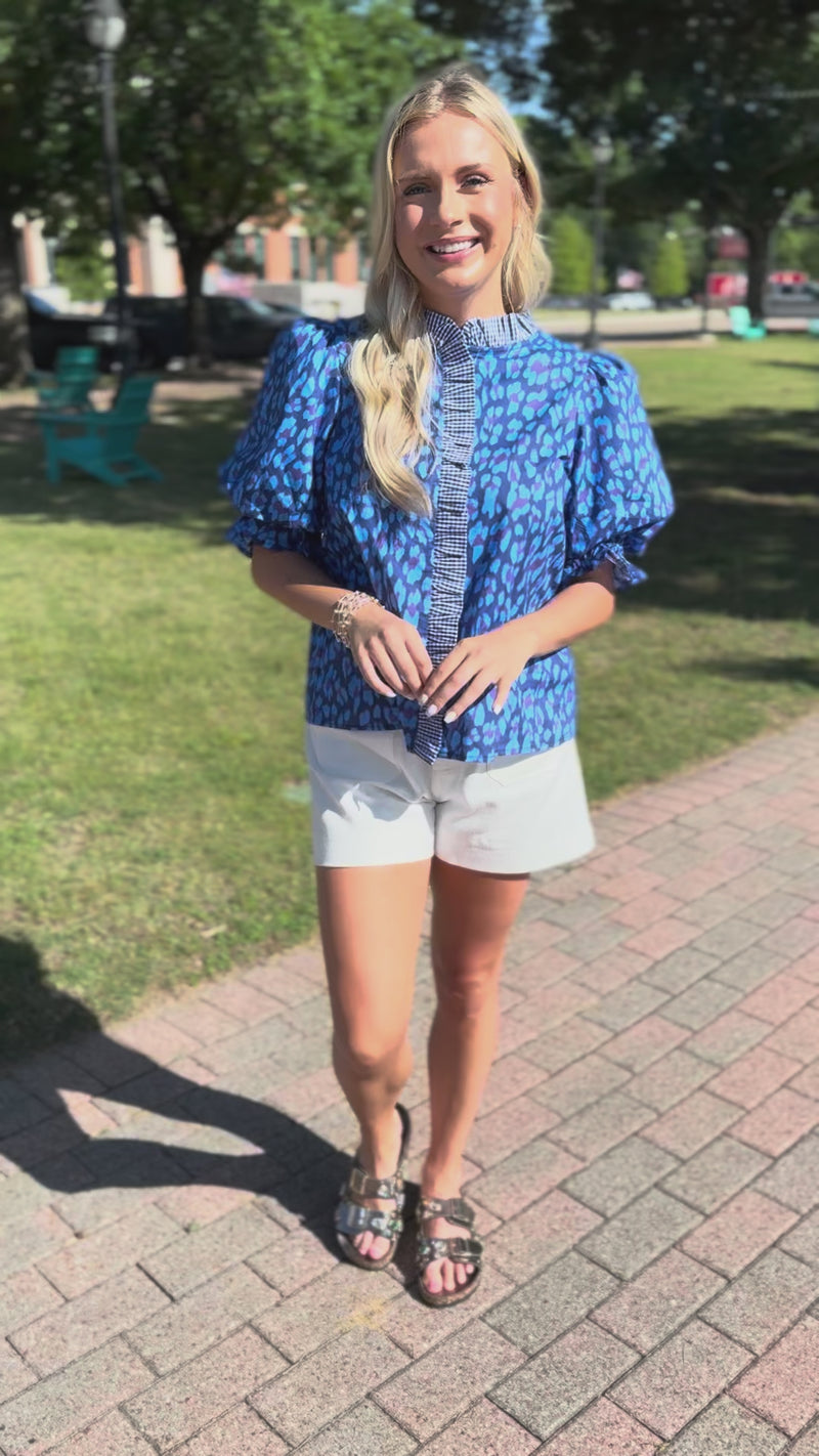 A woman modeling a blue and white patterned puff-sleeve blouse with a buttoned placket, paired with white shorts and brown sandals, standing outdoors.