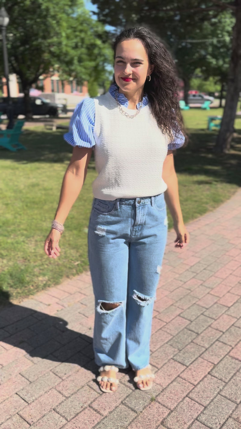 A woman walking outdoors on a sidewalk, wearing a white sleeveless top over light blue and white gingham short-sleeve blouse, light-wash distressed jeans, and white slide sandals.