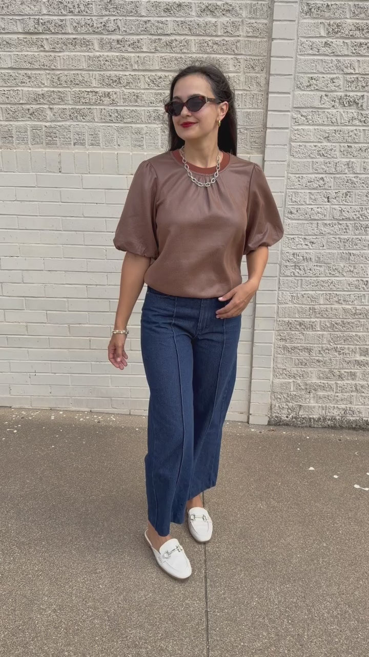 Brunette model standing outside wearing a brown blouse and denim jeans wearing black sunglasses.