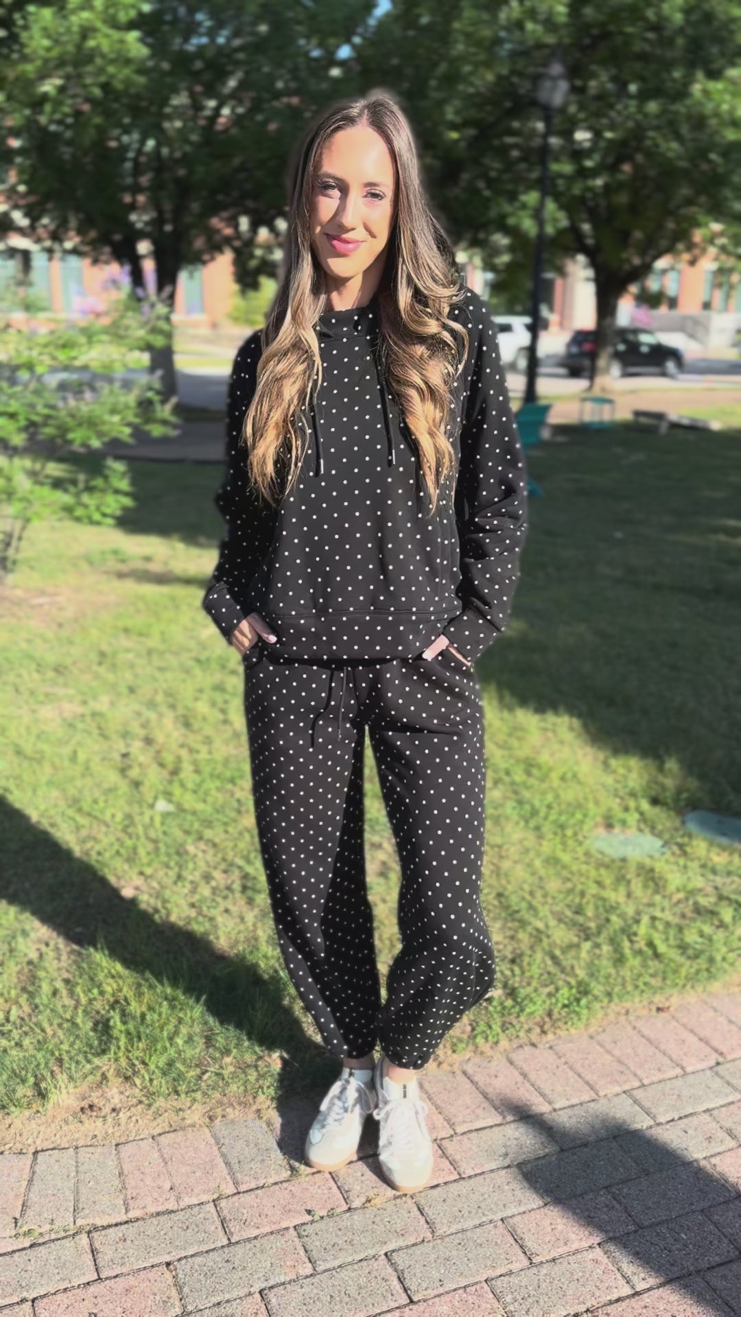 A woman modeling a matching black and white polka dot lounge set, consisting of a long-sleeve top and jogger pants, standing on a concrete sidewalk