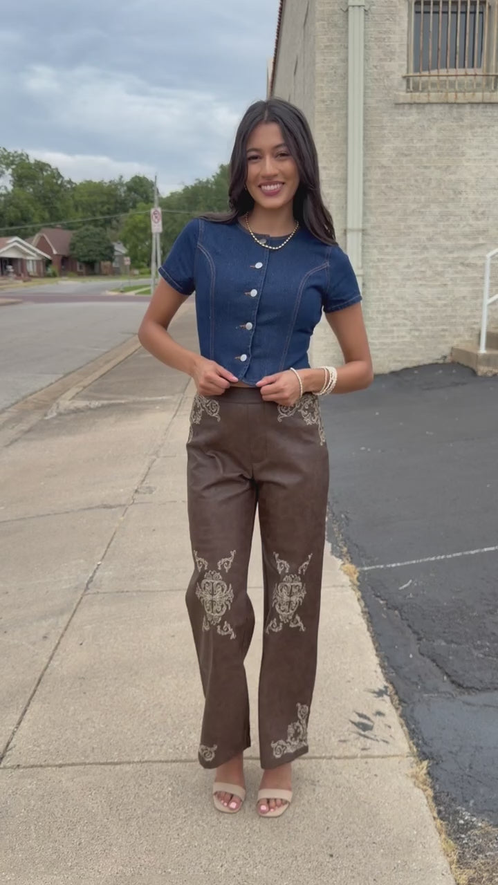 Brunette model wearing a denim top with buttons and a faux leather brown pant standing outside. Southern House Clothing