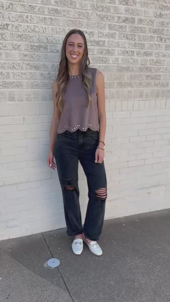 A woman wearing a sleeveless dusty mauve top with scalloped studded trim and dark jeans, standing outdoors in front of a building.