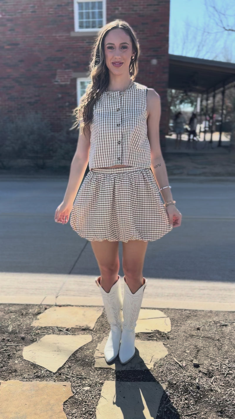 Woman wearing a checkered tank and skirt and white boots standing in front of a building.