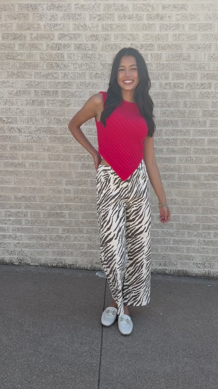 A woman posing outdoors on a sidewalk next to a light brick wall, wearing a bright coral-red ribbed knit sleeveless top and high-waisted, wide-leg zebra-print pants in beige and brown, paired with white slide sandals.