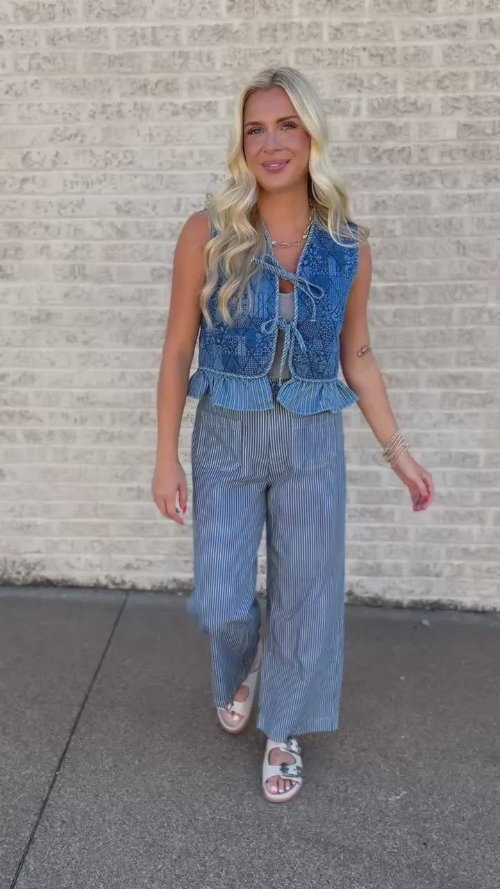 A smiling blonde woman stands outdoors on a sidewalk next to a light brick wall, wearing a sleeveless blue patterned vest with a tie-front detail over a gray top and blue-and-white pinstriped pants.