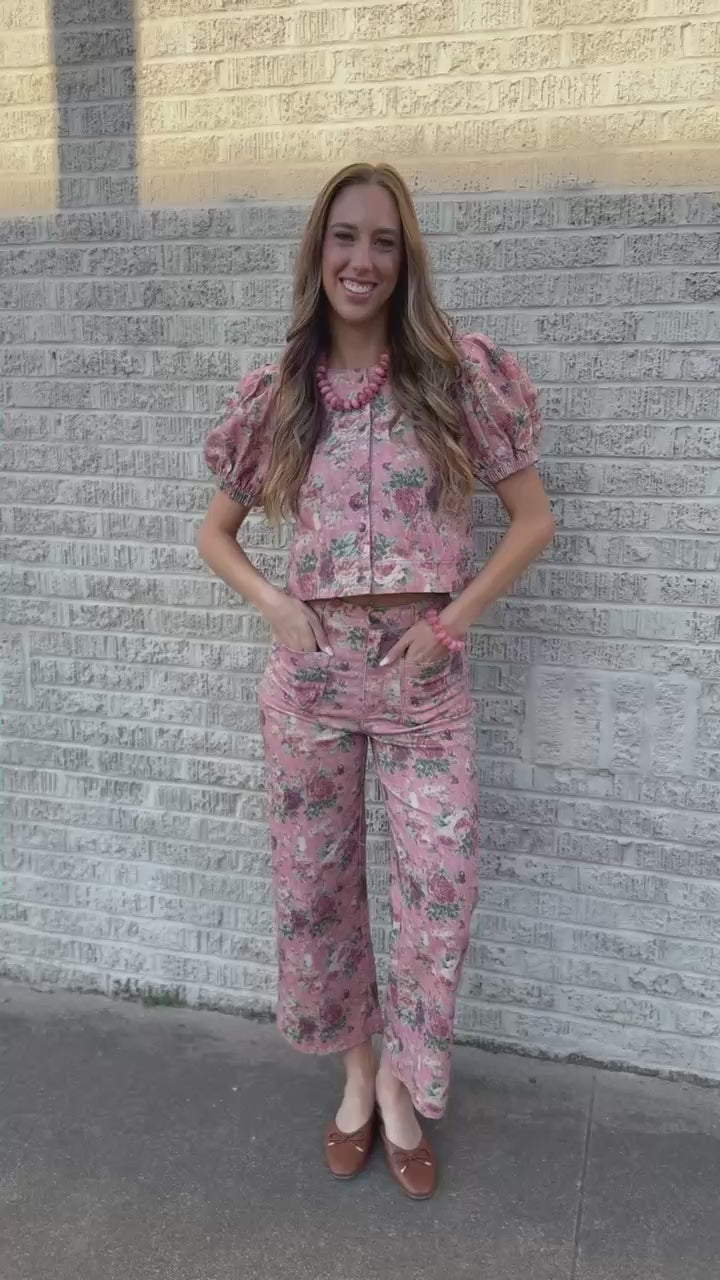A woman wearing a pink floral puff-sleeve button-up shirt and matching floral pants, standing outdoors against a light neutral wall.
