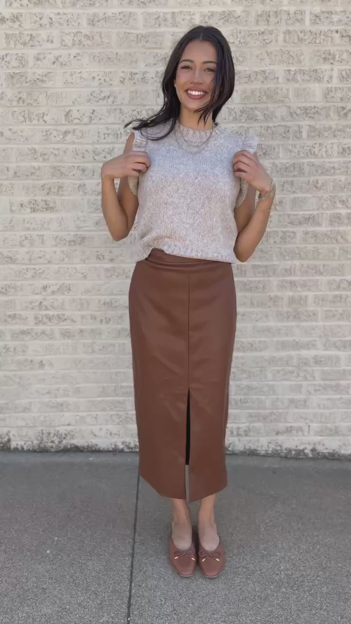 Model wearing a oatmeal colored sweater top with a cognac colored faux leather skirt standing outside.
