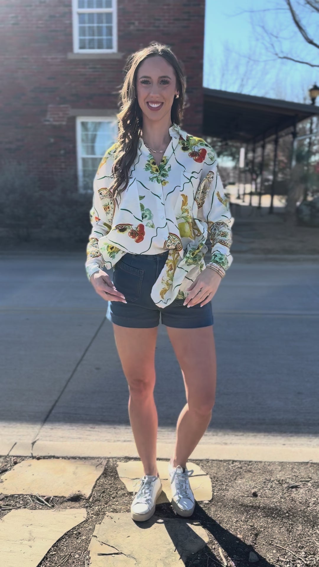 Woman wearing a floral shirt and blue shorts standing outside.