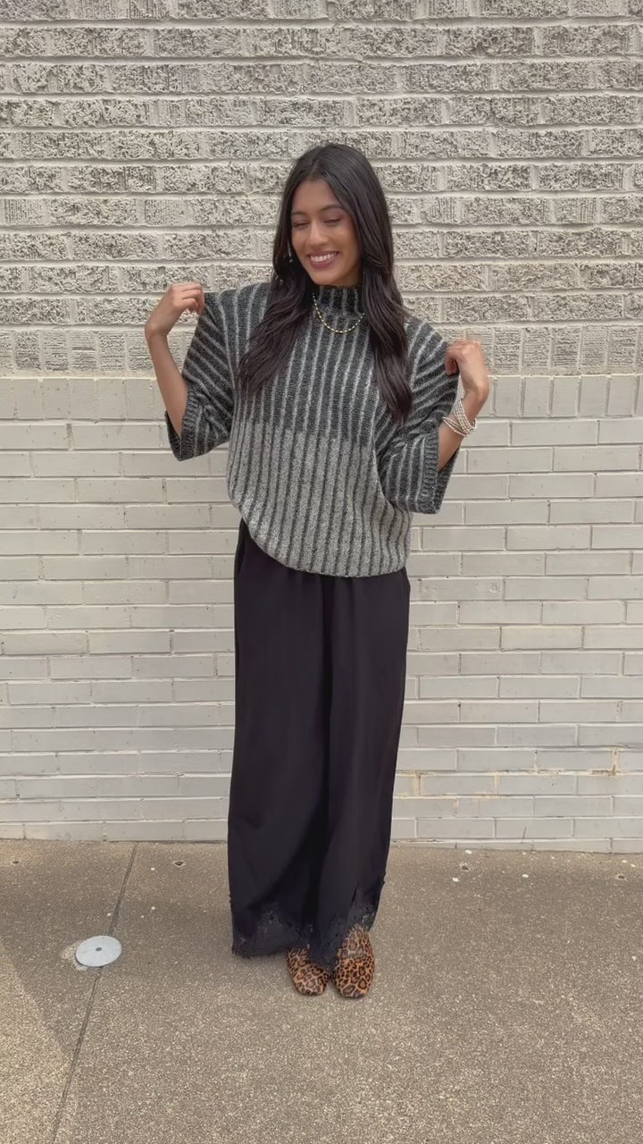 Brunette model wearing a black sweater top and black pants with a lace detail standing outside. Southern House Clothing