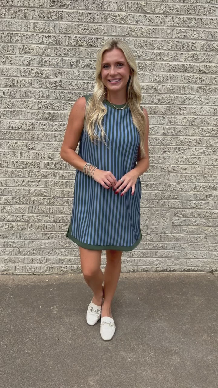 Blonde model wearing a green and blue striped dress and white flats standing outside. Southern House Clothing. Dress name is called the Aledo Lane Striped Dress