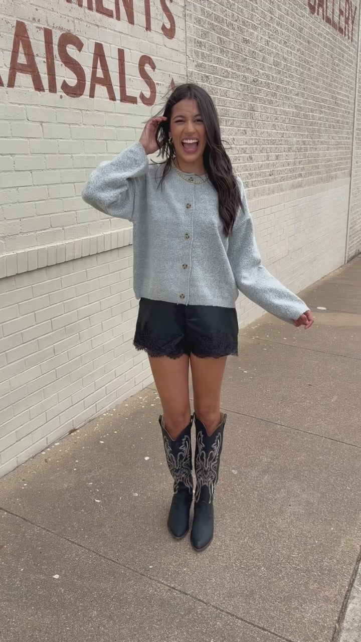 Brunette model standing outside wearing the Ashford Everyday Knit Cardigan in Heather Gray featuring a soft textured knit, round neckline, tortoiseshell-style buttons, and relaxed fit, paired with black lace-trim shorts and the Kevin cowboy boots in black. Southern House Clothing