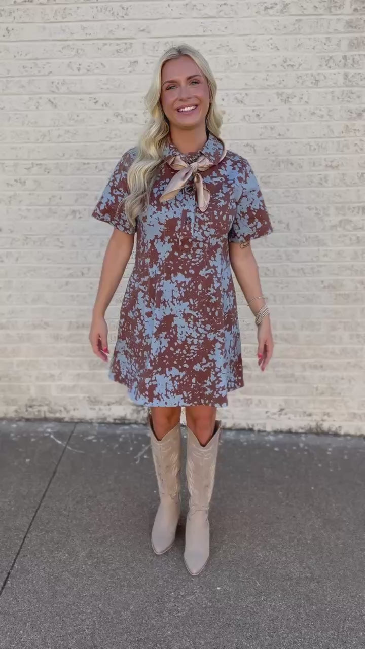 A woman stands on a sidewalk wearing a short-sleeve brown and light blue floral dress with a beige bow tie at the collar, paired with beige cowboy boots.