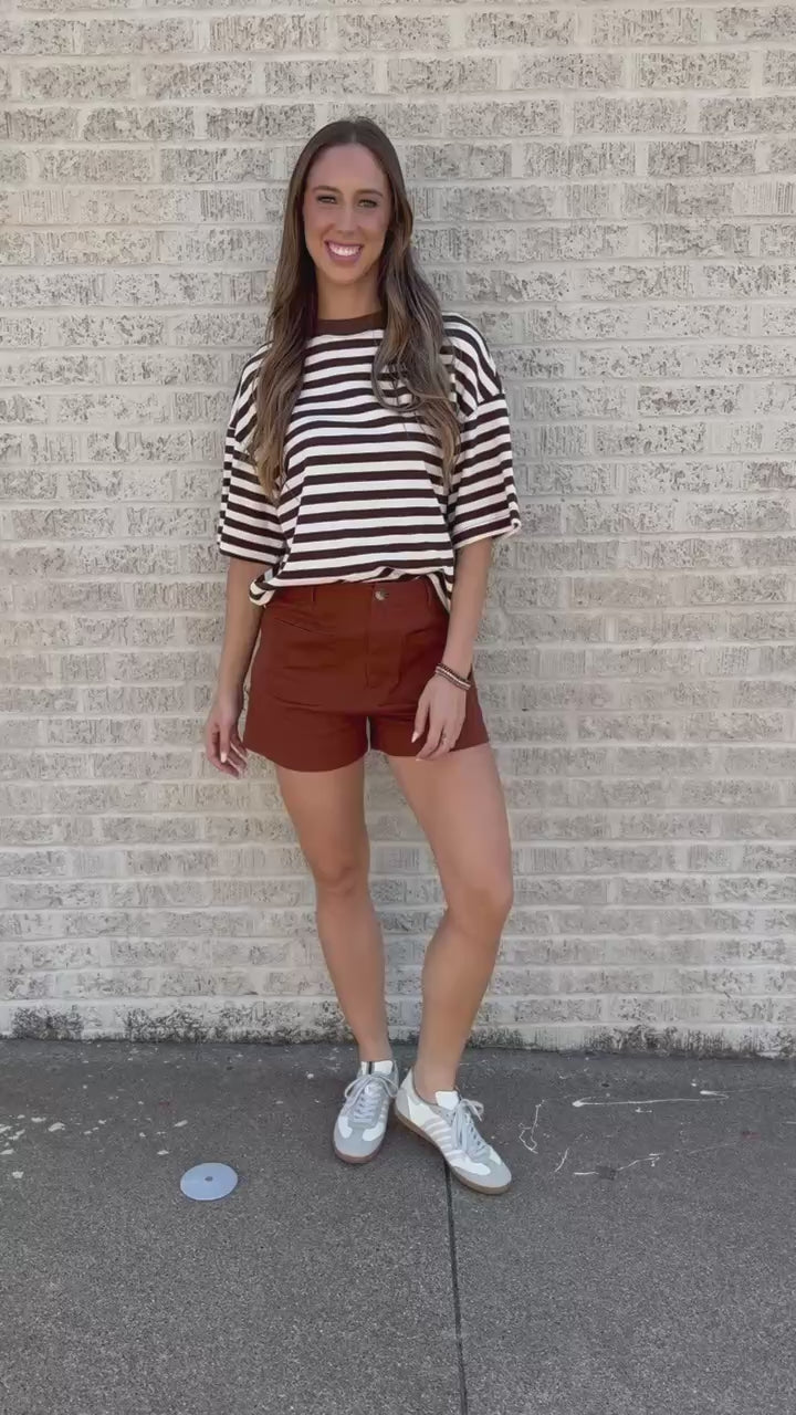 A woman posing outdoors against a light brick wall, wearing a brown and white striped t-shirt, rust-colored shorts, and white sneakers.