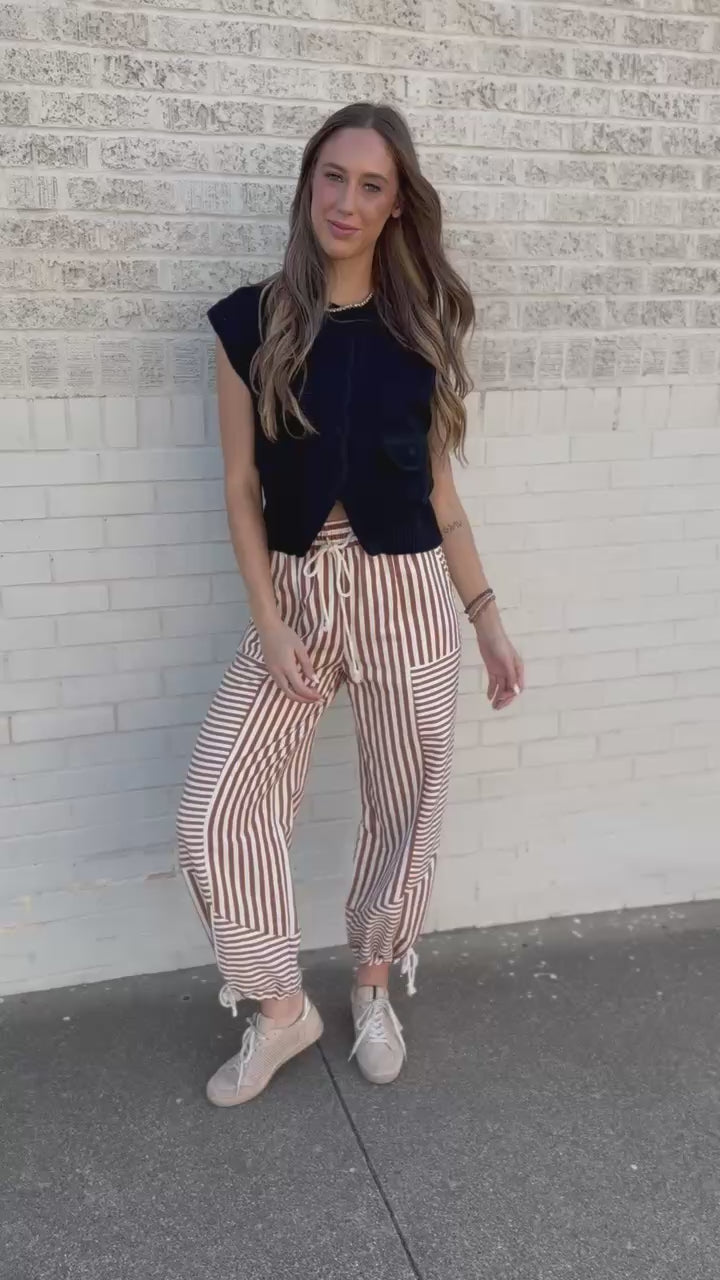 A woman wearing a black sleeveless button-front knit vest and rust-and-white vertically striped drawstring pants, standing outdoors in front of a building 