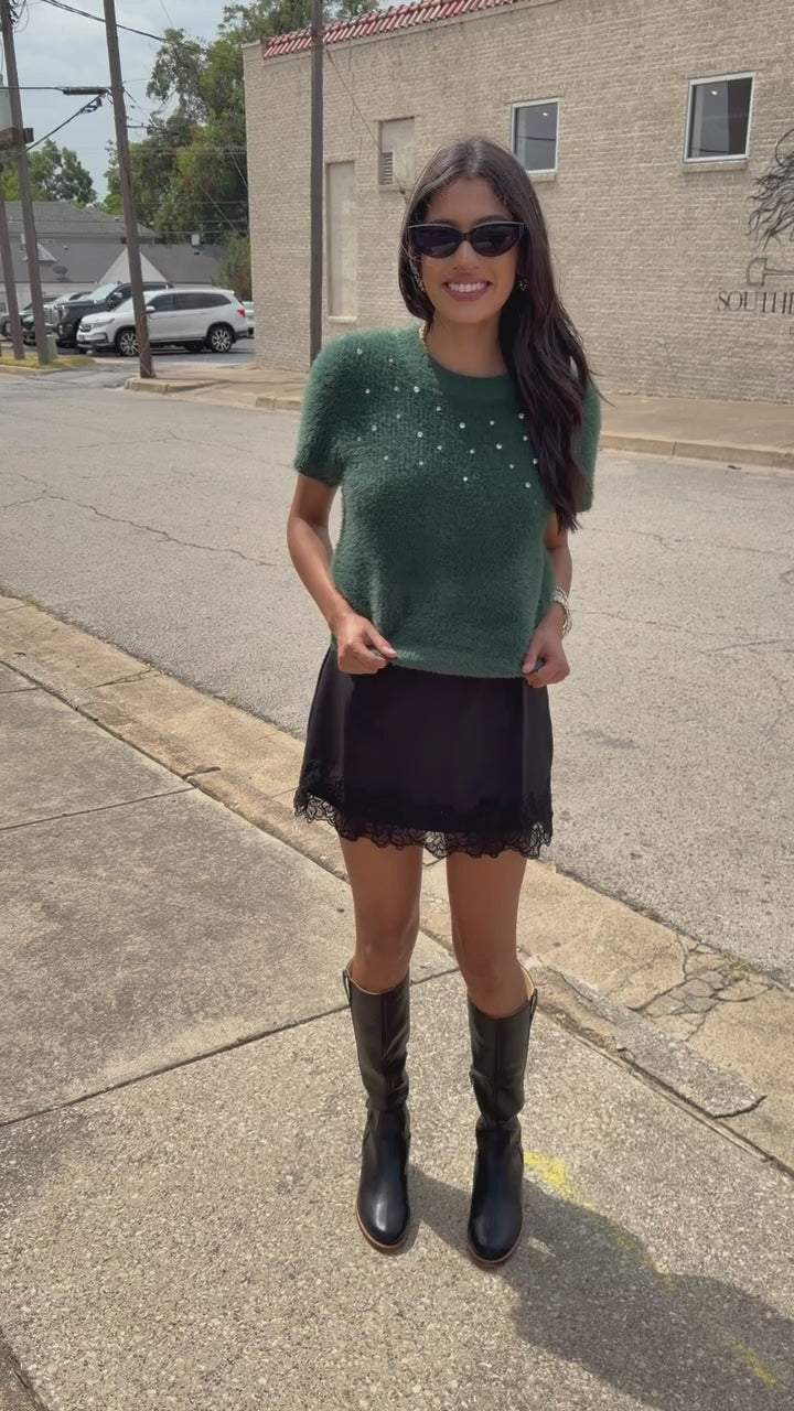 A brunnette model wearing the Liora Pearl Knit Top – Hunter Green, the Bellarosa skort in black and the harriett tall riding boots in black standing outside. Southern House Clothing