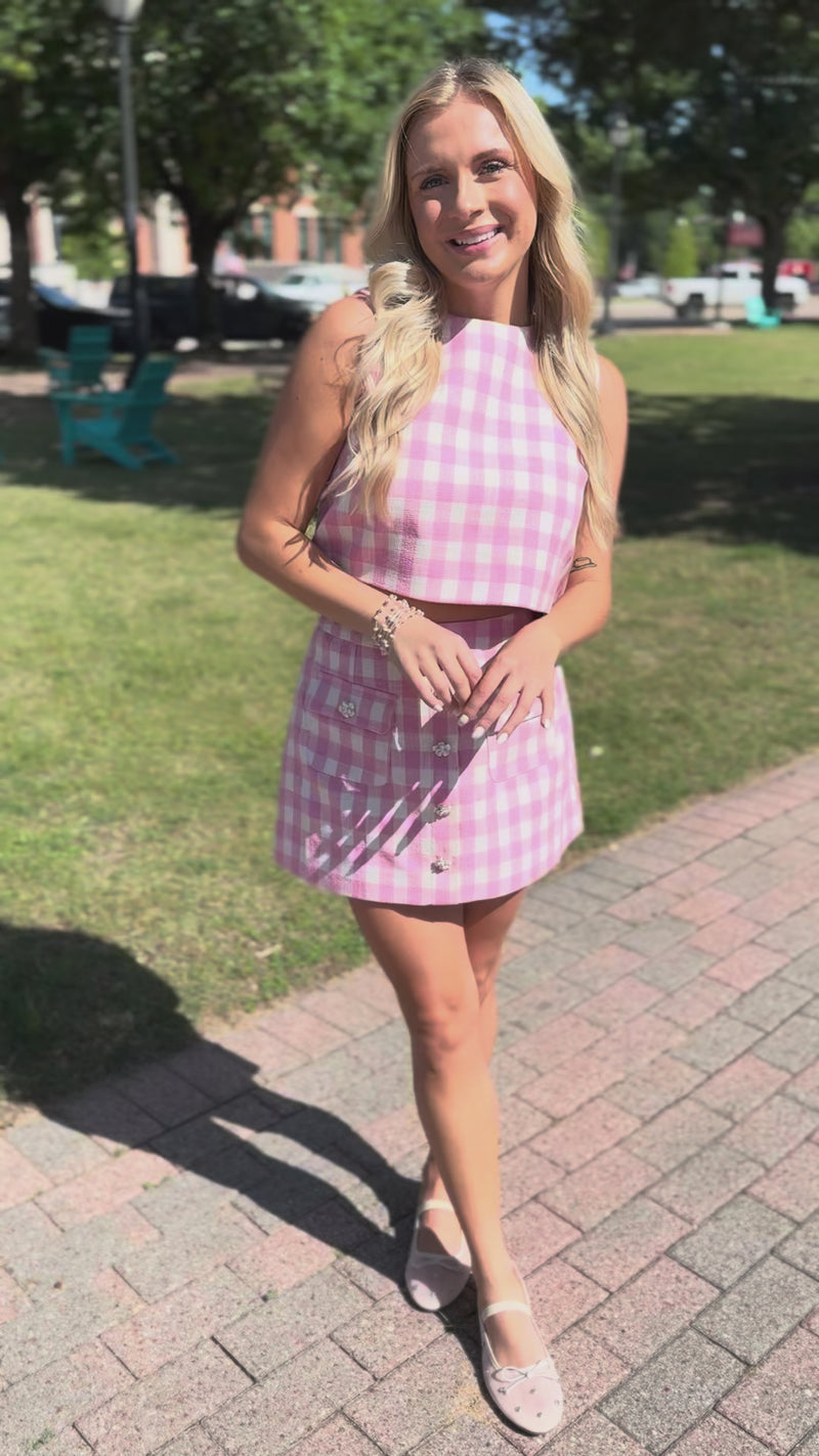Blonde model wearing a pink and white checkered tank and skirt set outside.