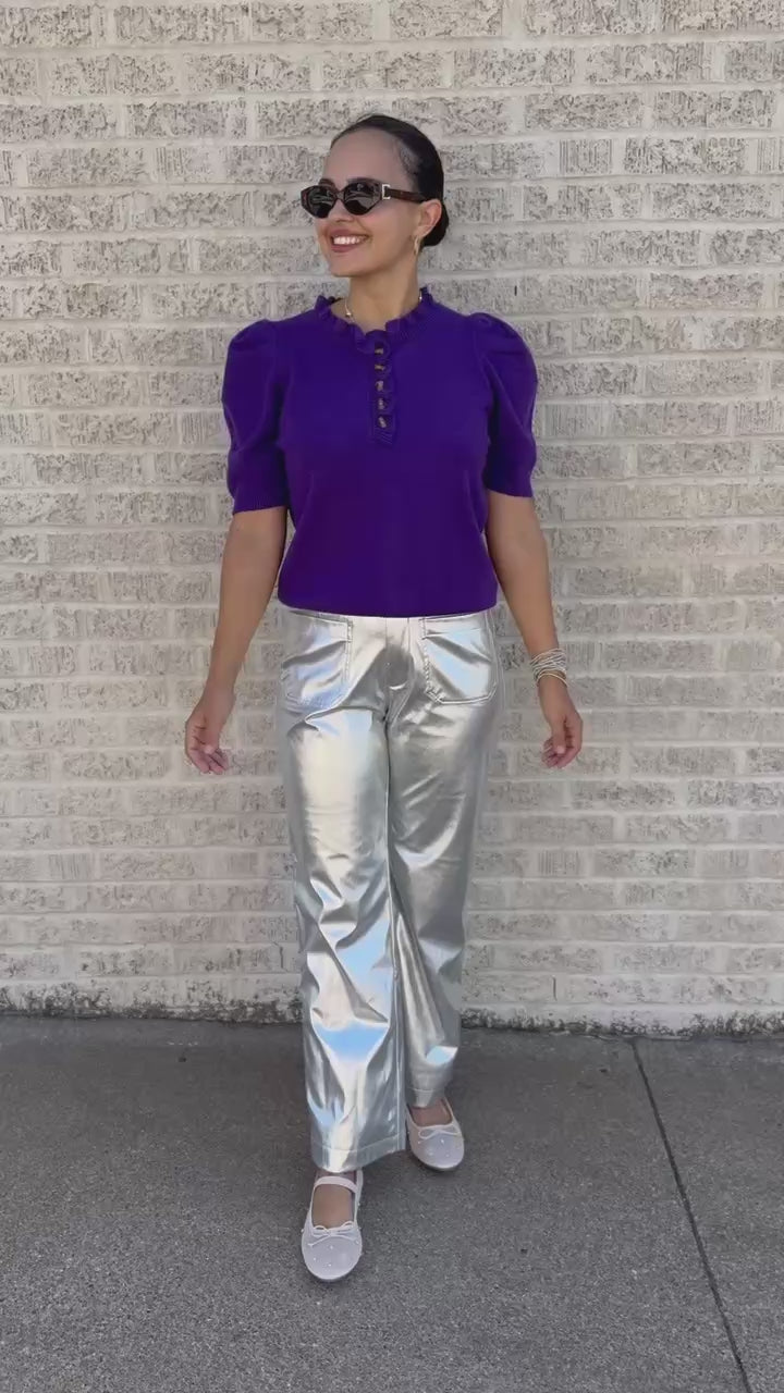 A woman wearing a purple short-sleeve knit top with puff sleeves and a buttoned ruffled neckline, paired with shiny metallic silver pants, walking outdoors next to a light brick wall.