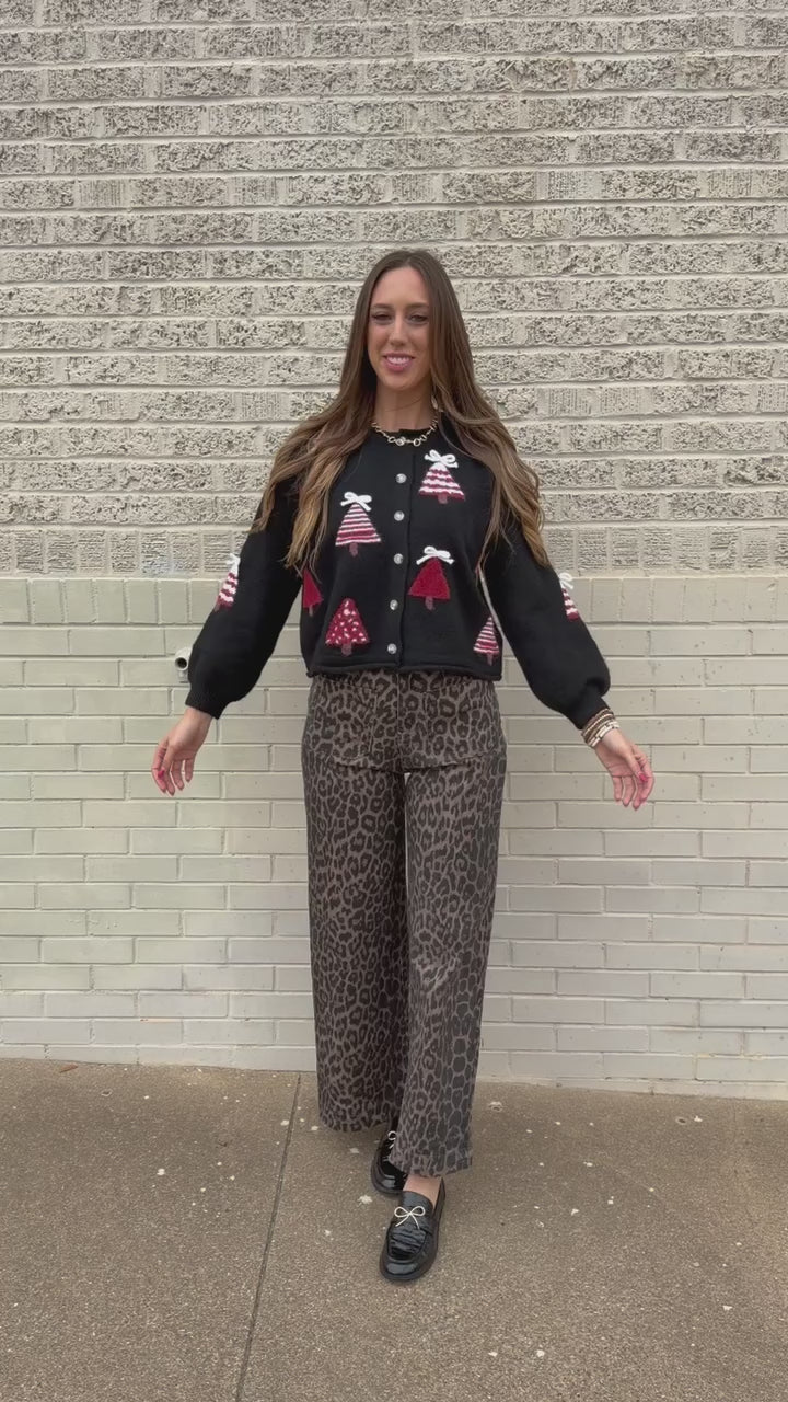 Blonde model standing outside wearing Merry Little Christmas Tree Cardigan in black featuring red and white Christmas tree designs, dimensional white bows, and front button closures, styled with Wildwood Leopard Pants. Southern House Clothing
