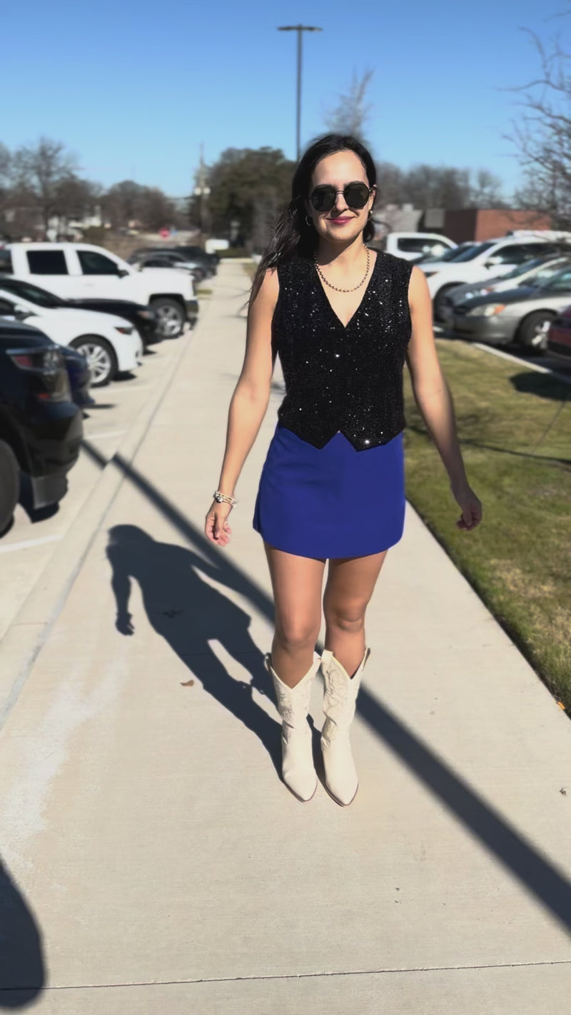 Woman wearing a black sequin top, blue skirt, and white cowboy boots outside.