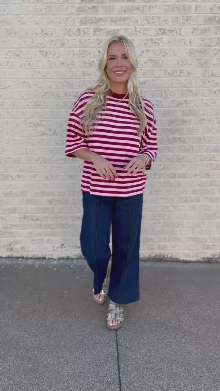 A woman stands outdoors against a light brick wall wearing a red and white horizontally striped short-sleeve top, dark navy wide-leg pants, and patterned sandals.