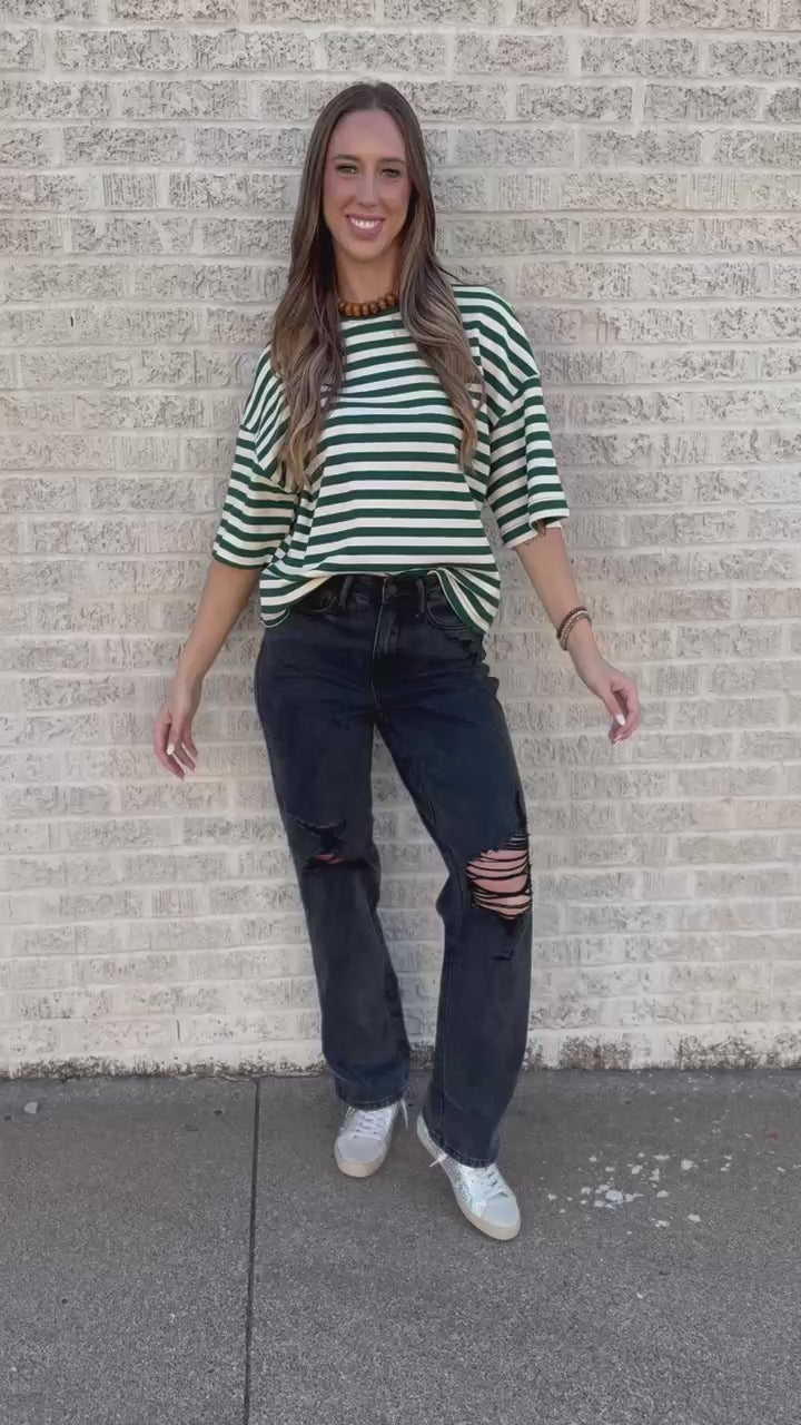 A woman stands on a sidewalk wearing a green and white striped short-sleeve top, black distressed jeans, and white sneakers.