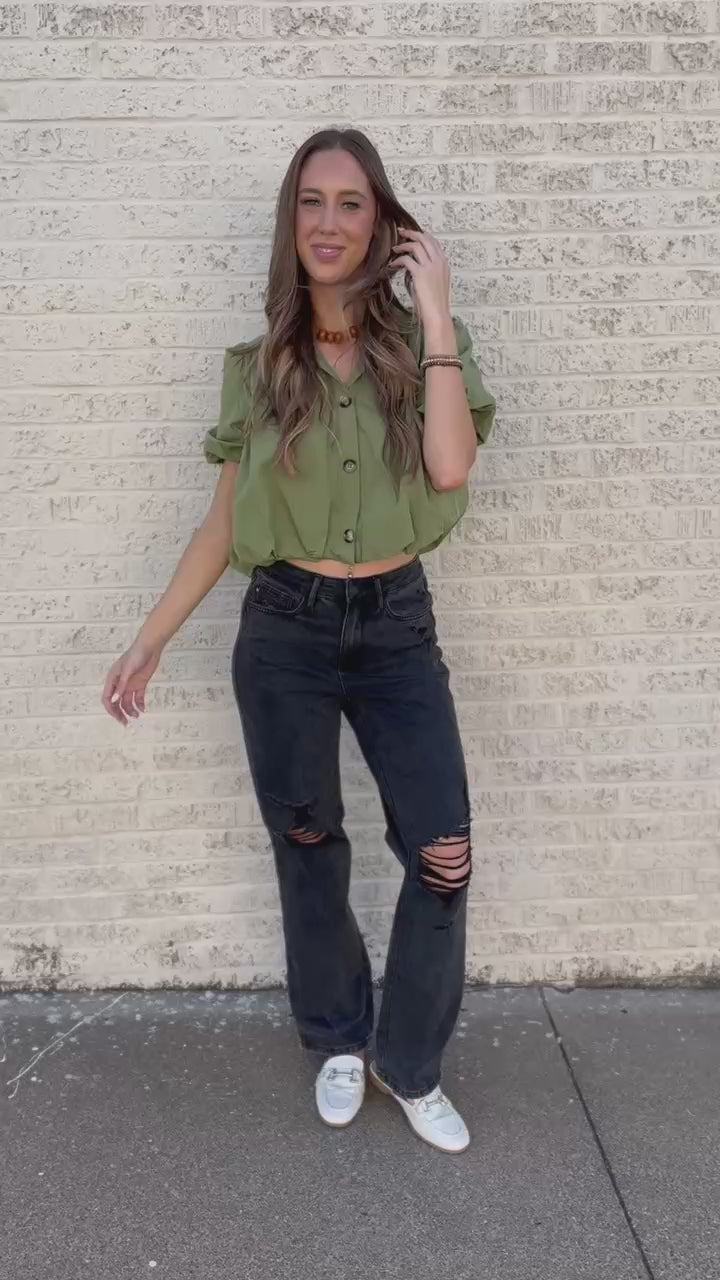 A woman stands against a light beige brick wall wearing an olive green short-sleeve button-up shirt, black distressed jeans, white loafers