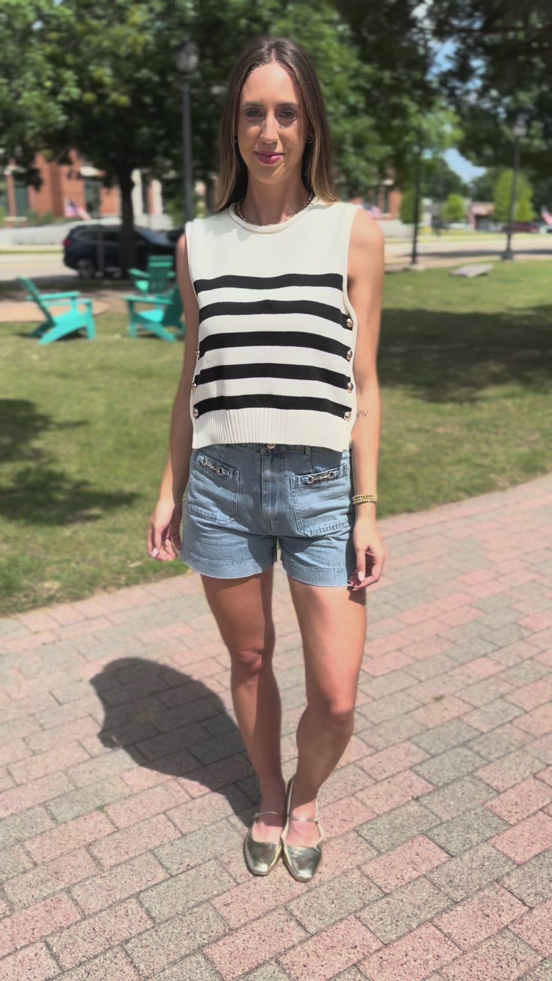 A woman wearing a sleeveless knit top with bold black and white horizontal stripes, accented by gold-tone buttons down the side, paired with denim shorts in an outdoor setting.
