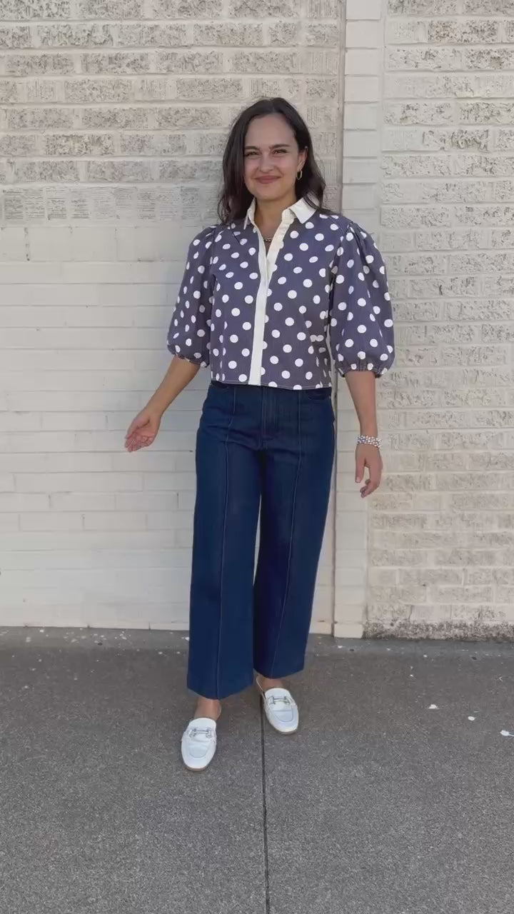 A woman wearing a purple and white polka dot blouse with dark blue jeans, standing against a light-colored brick wall.