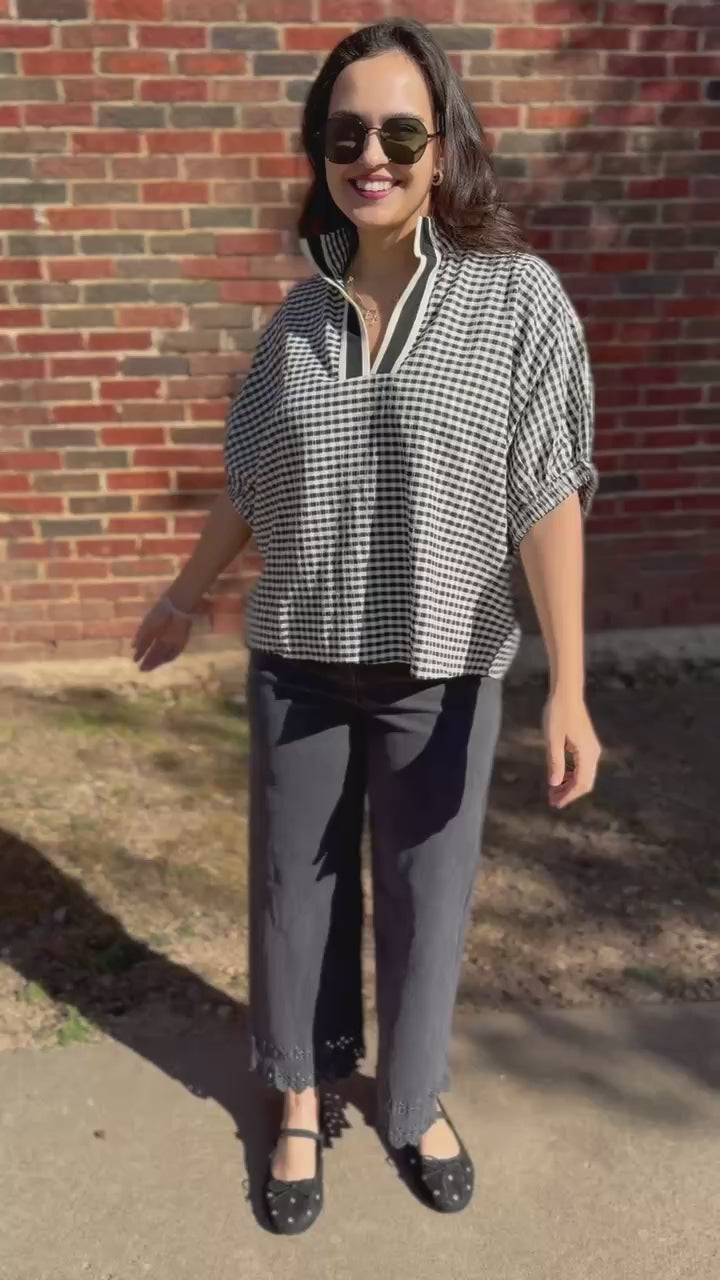 Woman wearing a checkered blouse and dark jeans standing in front of a brick wall 