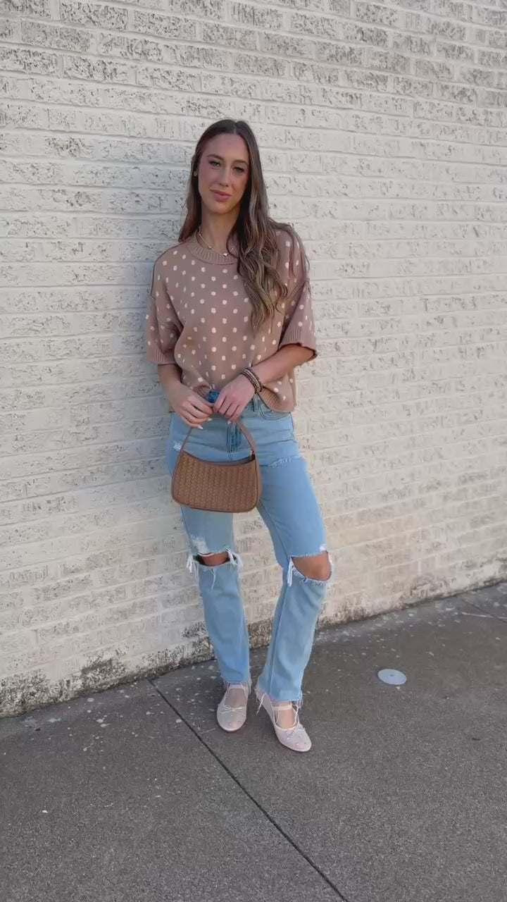 Blonde model standing outside near a brick wall wearing a camel colored sweater top with off white dots, blue jeans and flats.