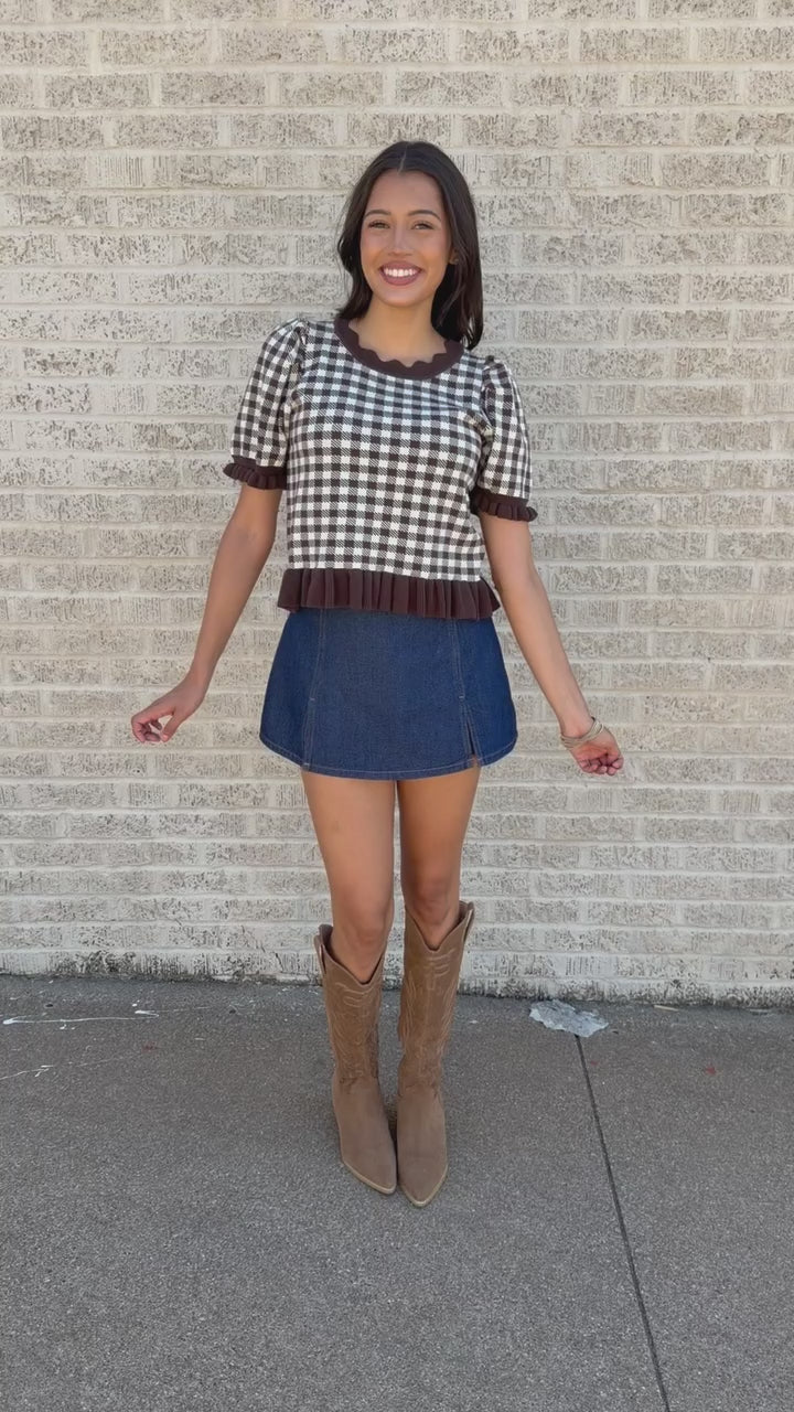 A woman stands on a sidewalk wearing a black and white gingham top with dark brown ruffle trim, a blue denim mini skirt, and brown cowboy boots.