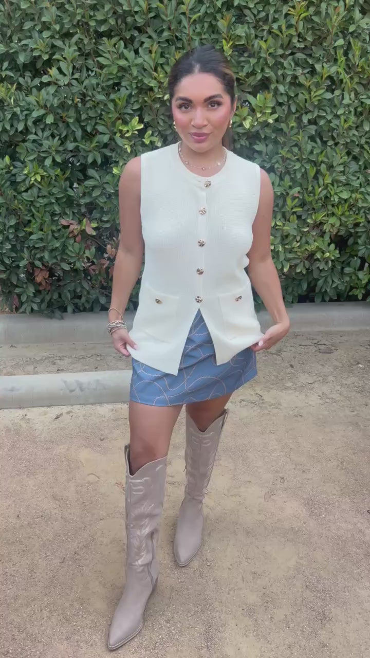 Brunette model in a cream vest and a blue skirt in an outdoor setting. Southern House Clothing