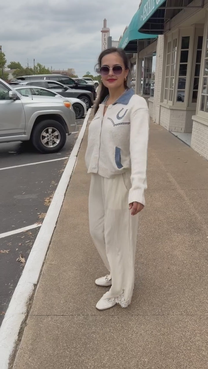 Brunette model wearing an Oatmeal knit Pecos Bridle Embroidered Cardigan with dusty blue collar, horseshoe embroidery, and front buttons, styled with cream trousers. Southern House Clothing